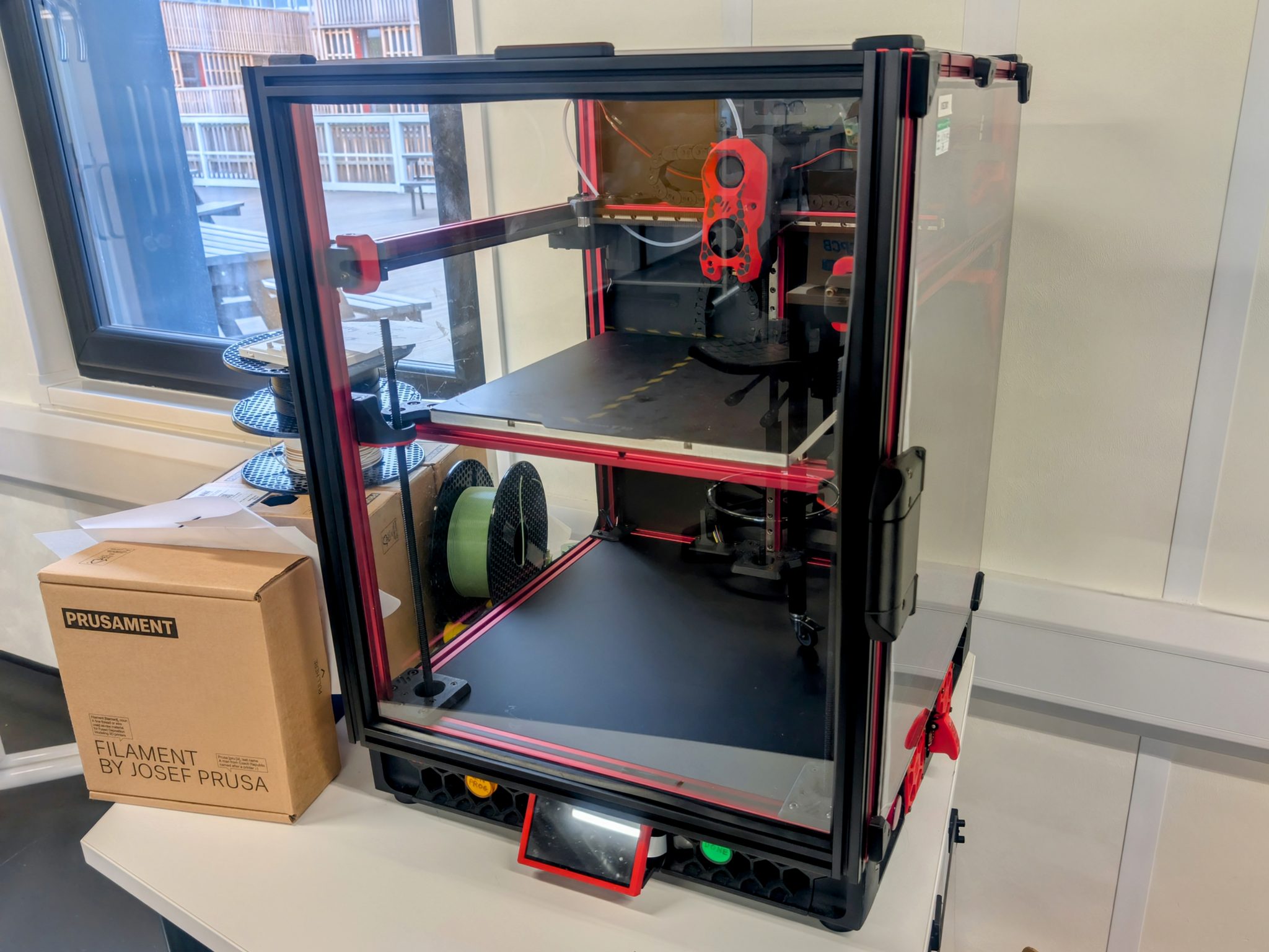 A red and black 3D printer next to filament spool boxes